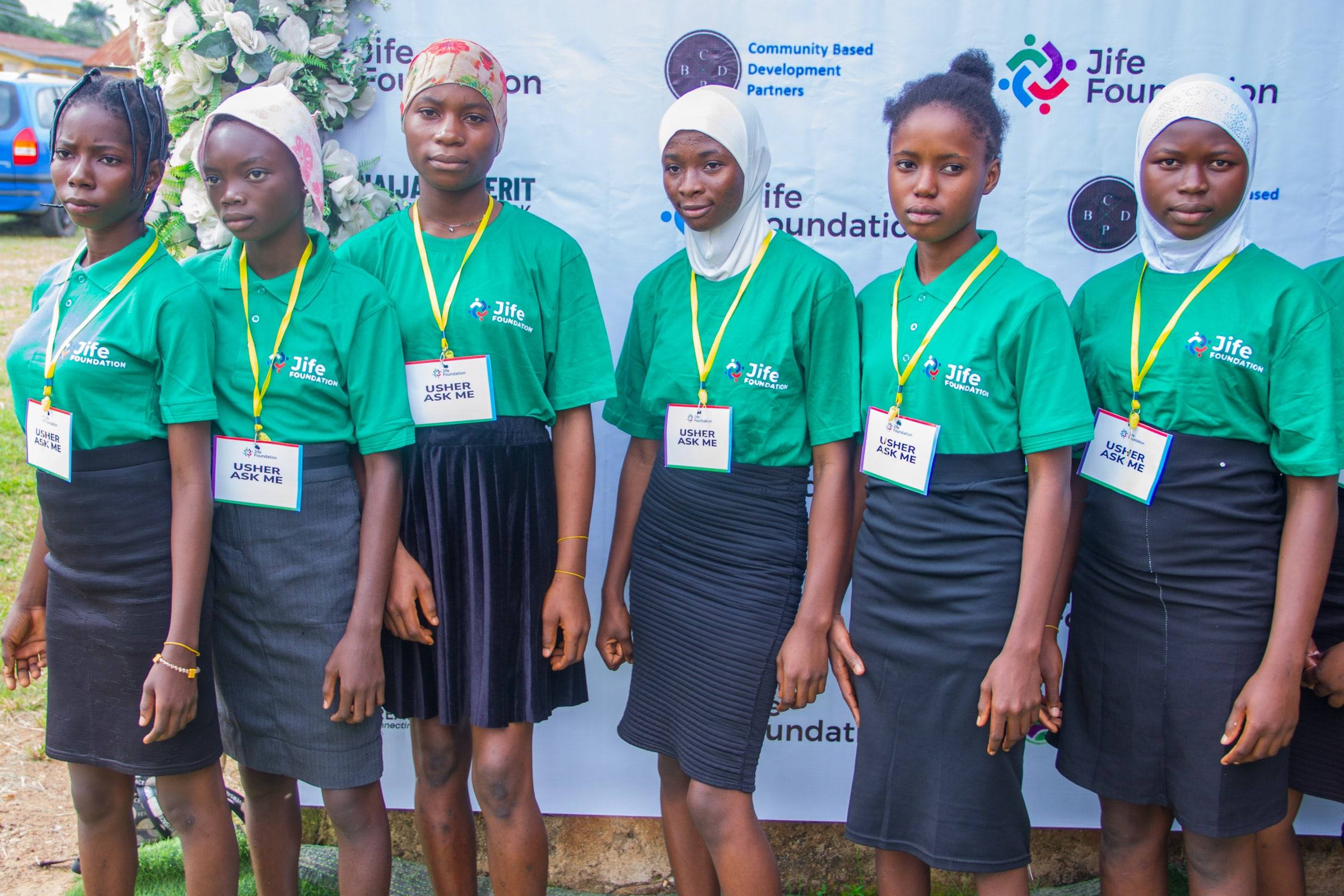 Five young women in JIFE Foundation T-shirts wearing lanyards marked Usher, standing in front of an event backdrop.