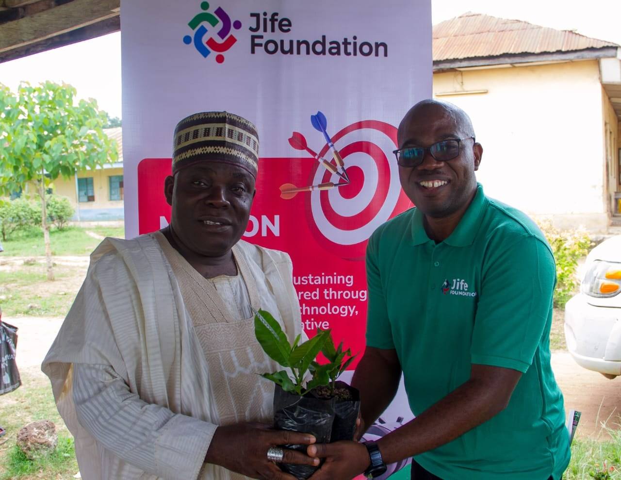 A JIFE Foundation team member handing a potted cashew seedling to a community elder in front of a Jife Foundation banner.