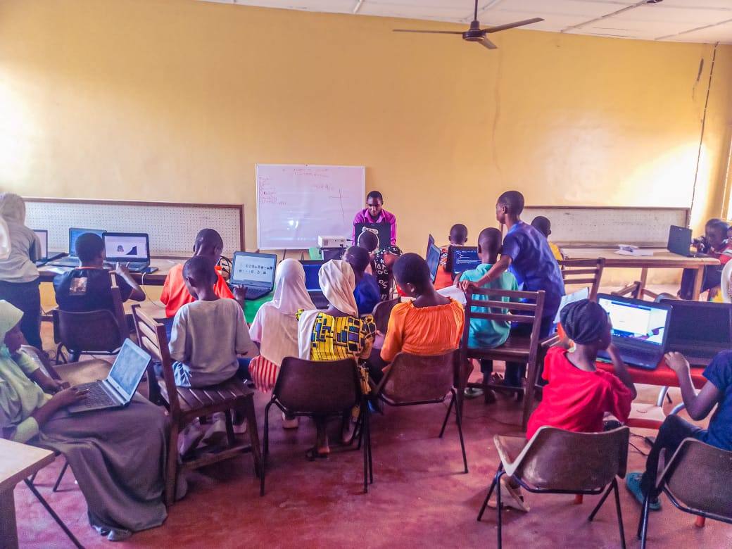 Children seated at laptops in a classroom in Kwara State, with an instructor standing at a whiteboard at the front.