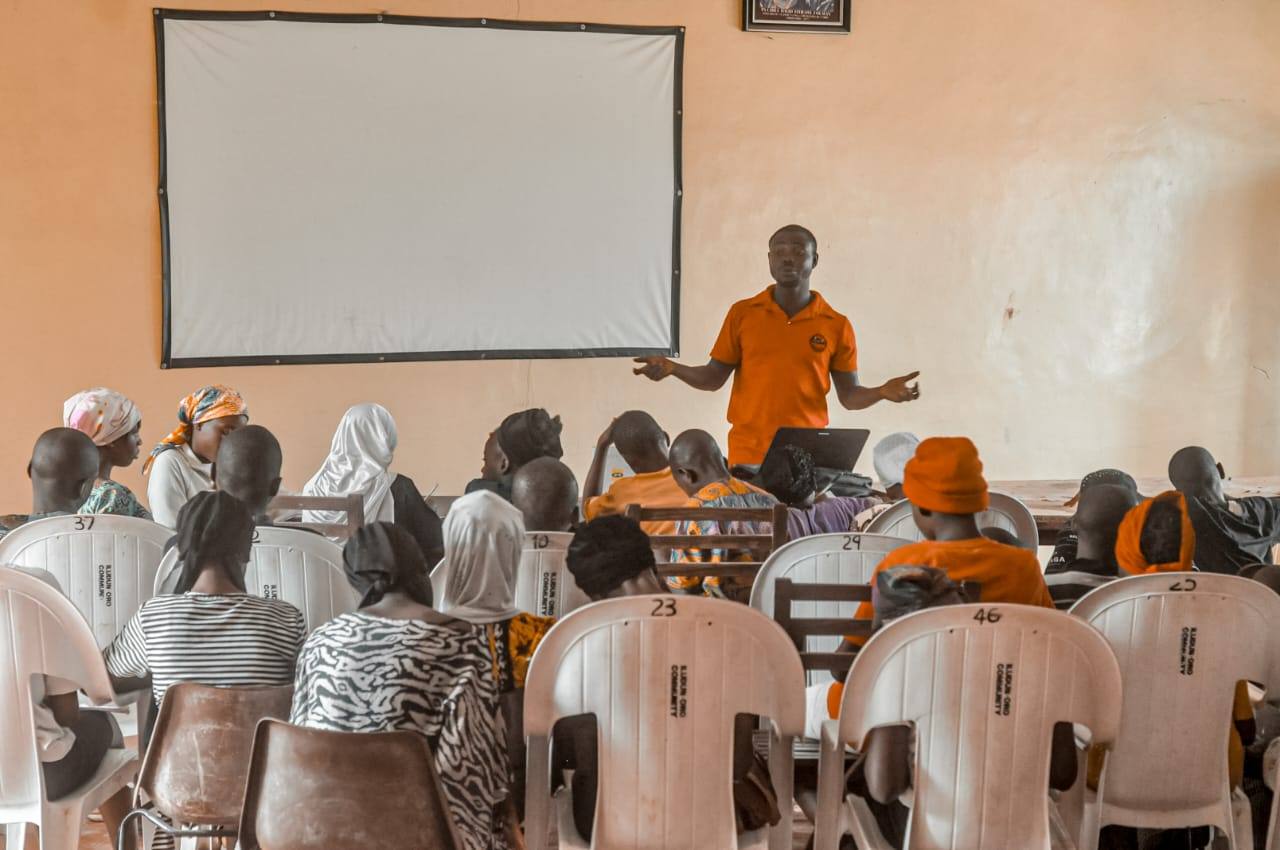 An instructor in an orange JIFE polo shirt addressing a seated group of young people in a classroom, with a projector screen behind him.